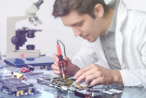 Electronics background. Young energetic male tech or engineer repairs electronic equipment in research facility. Shallow DOF, focus on the hands of the worker. This image is toned.